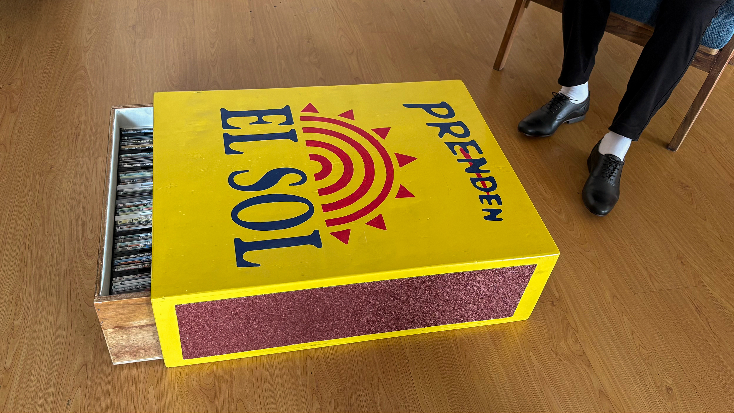 Installation view of Factory of Copies (2023), showing an enlarged hand-painted Ecuadorian matchbox with its drawer of pirated DVDs partially open beside a seated figure, indicating the work’s scale.