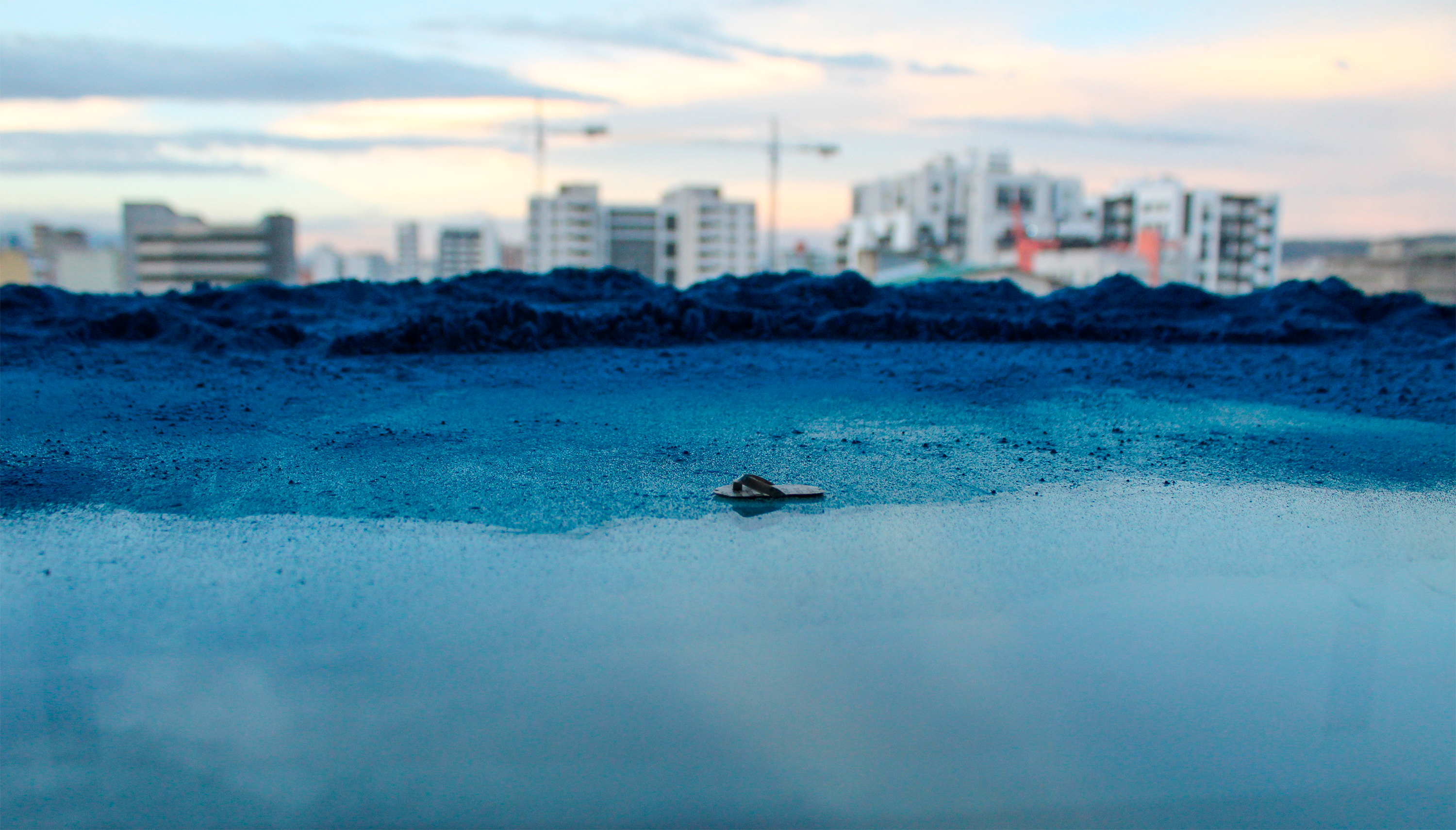 Installation view of La chancla perdida (2020), showing a miniature newspaper flip-flop on a glass surface covered with turquoise pigment, with the city visible in the distance.