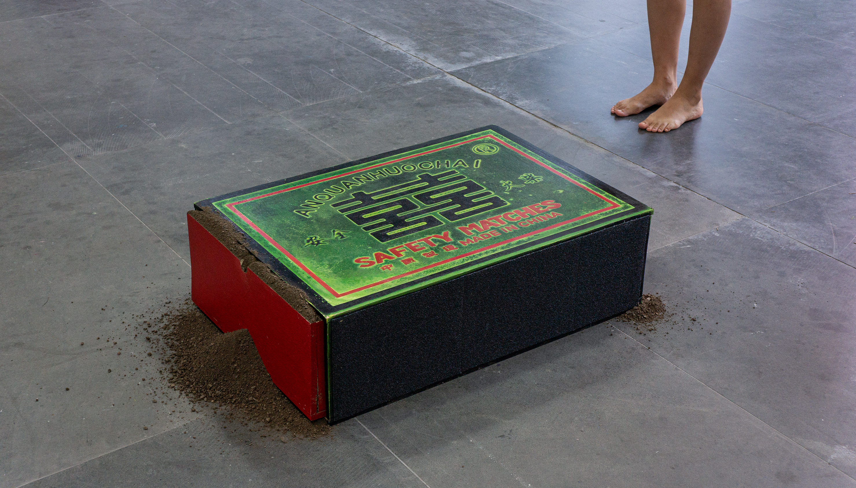 Installation view of The Matchbox Cannot Hold the Earth (2026): an enlarged Chinese matchbox filled with black soil, with a standing figure indicating its scale.