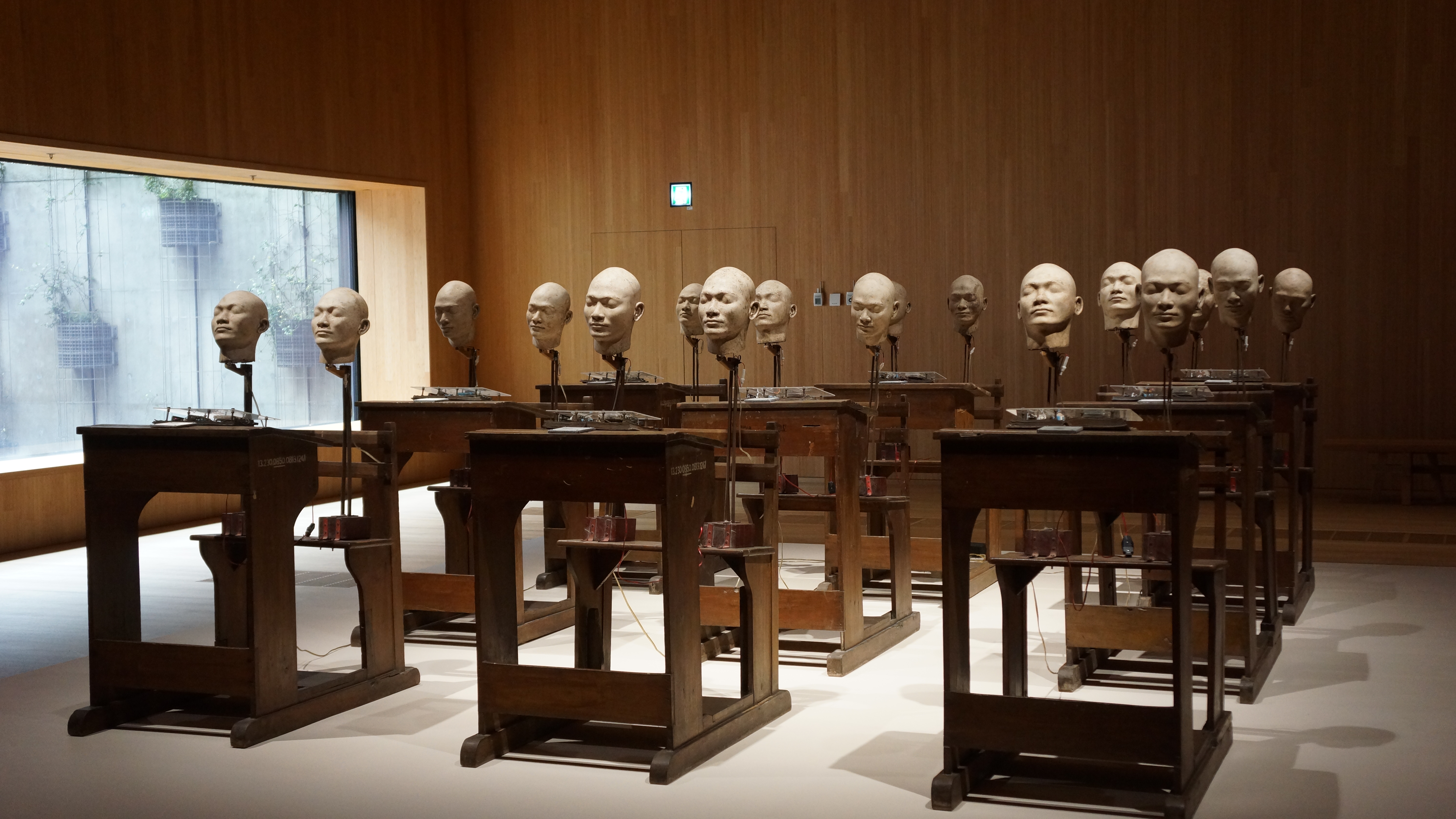 Instalación cinética "Fermentation of the Mind" de Heri Dono, con una fila de escritorios de madera antigua coronados por cabezas de fibra de vidrio blanca, en una sala de museo.